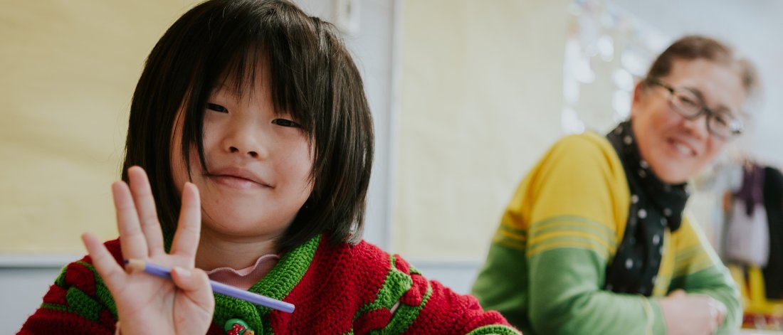 A child with long bangs and a smile holds up four fingers while drawing at a table.