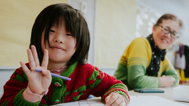 A child with long bangs and a smile holds up four fingers while drawing at a table.