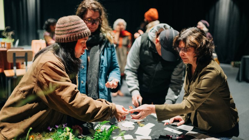A few people stand around a black table decorated with foliage. Two people with outstretched hands exchange an object.