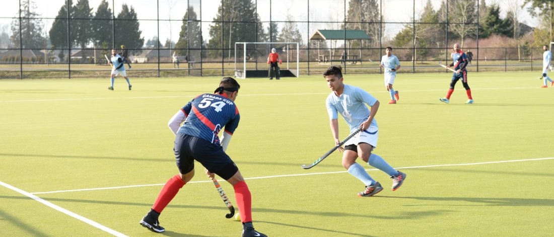 Field hockey players in action during a game on an outdoor turf field, with a goalie and other players in the background.