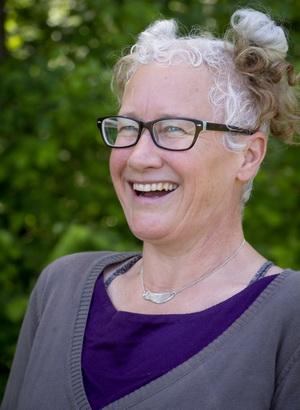 Photo of Tamara Unroe looking at the camera, a woman with glasses and white hair tied into two buns on top of her head, wearing a purple shirt and grew sweater