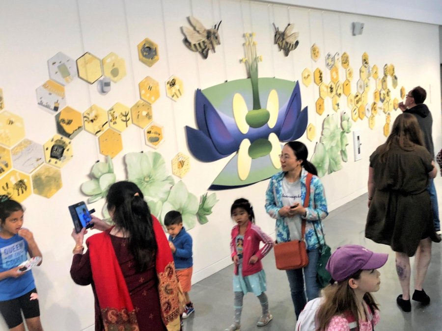 People viewing an interactive mural featuring bee-themed artwork on honeycomb panels at a community event.