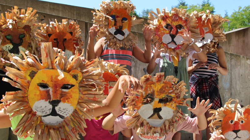 A group of kids stand and kneel in front of a concrete wall with handmade lion masks on their faces and arms out in claw pose.