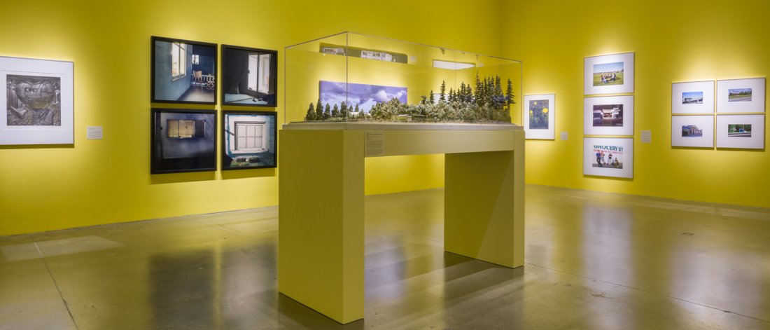 In a gallery space painted yellow hang multiple artworks on the walls. In the centre is a maquette of a Surrey residence in a vitrine mounted on a yellow wooden plinth.