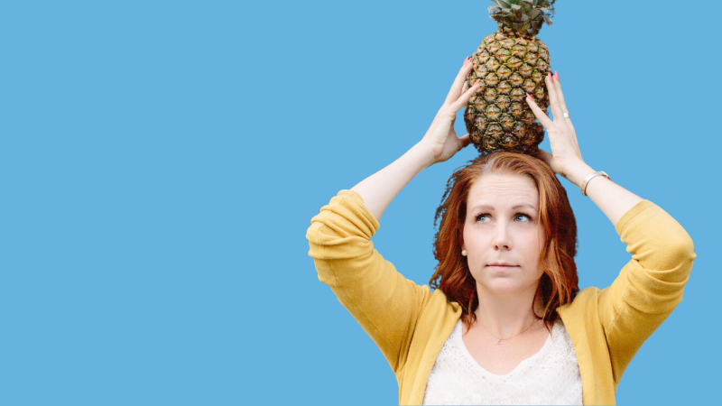 Katie-Ellen is wearing a yellow cardigan and holding a large pineapple on her head