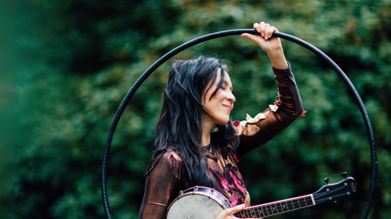 Performer Ginalina looks off camera as she stands in front of greenery while she holds her banjo and a hula hoop