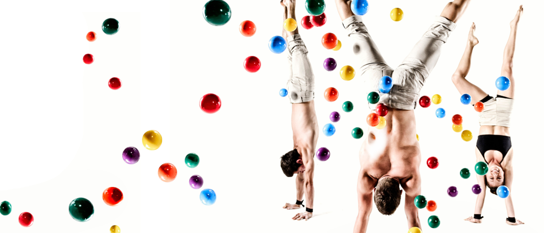 three acrobats doing handstands against a white background with 50 or more coloured balls bouncing all around them