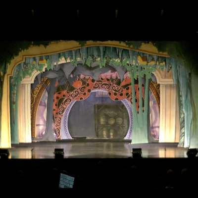 A photograph of a theatre stage that shows numerous backdrop structures, some green, some yellow, and some with flowers falling over the top. Some structures form a rectangular roof, while others are round portals. 
